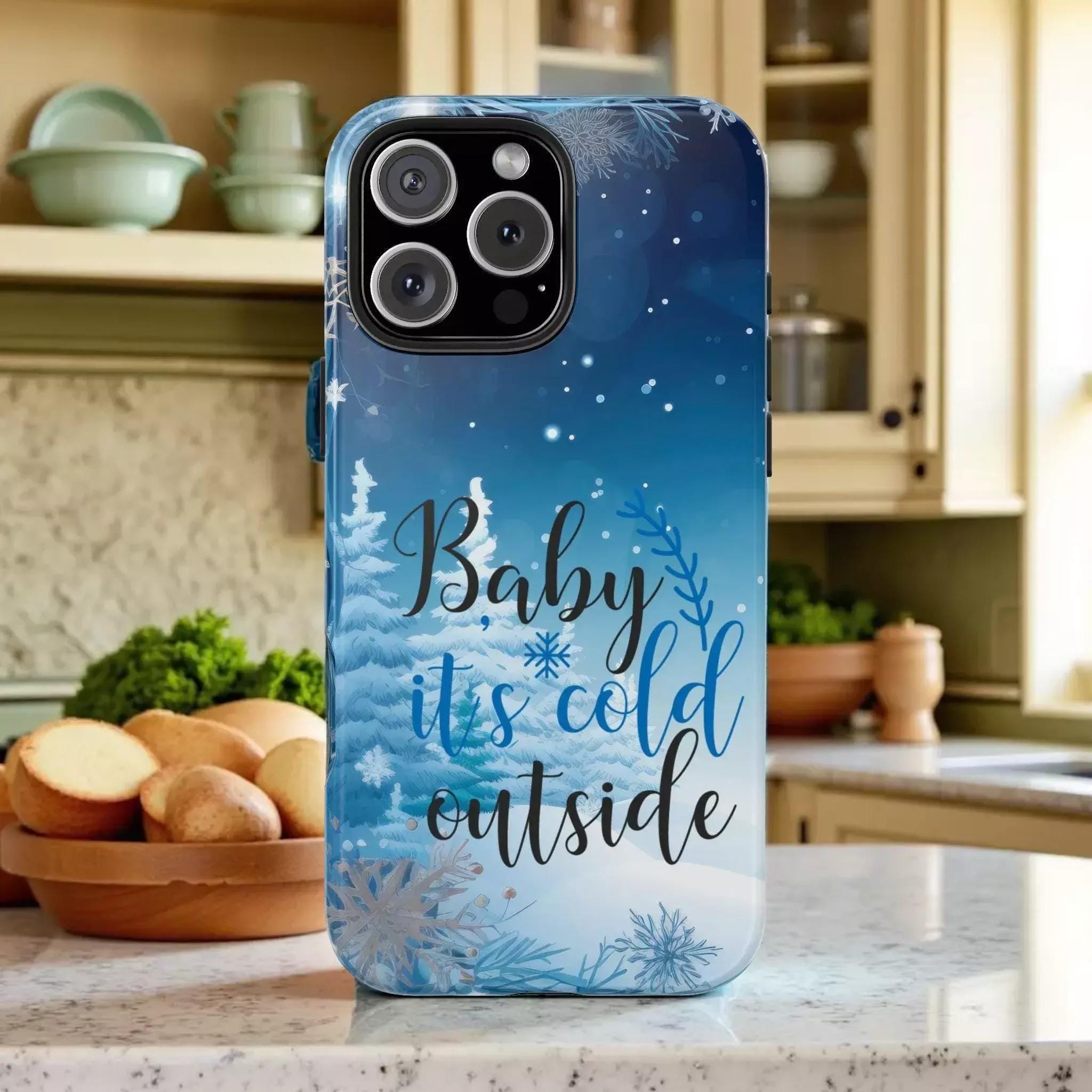 A phone case with a winter-themed design, featuring snowflakes and pine trees, along with the text "Baby it's cold outside".