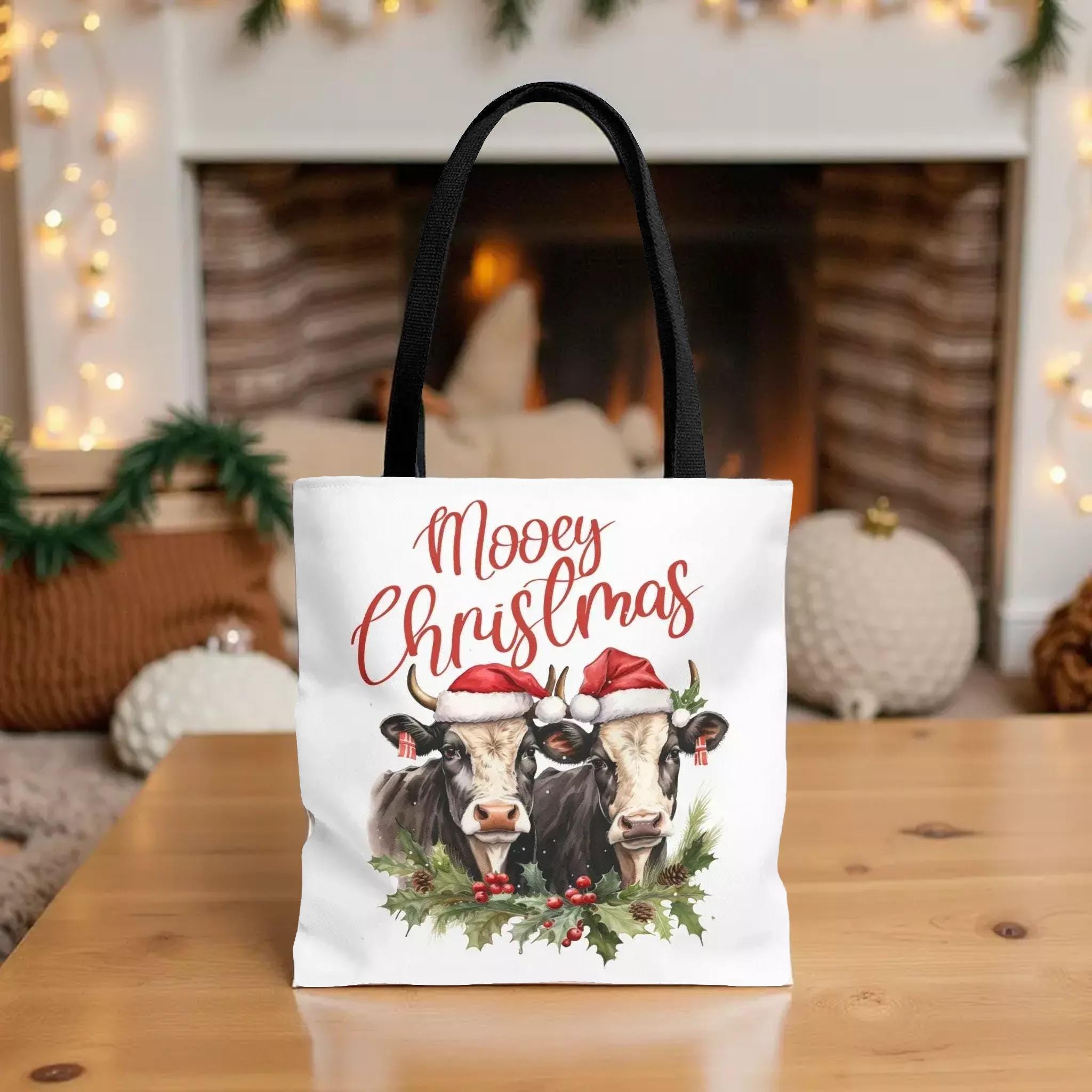 A white tote bag with a Christmas-themed design featuring two cows wearing Santa hats, surrounded by holly and berries.