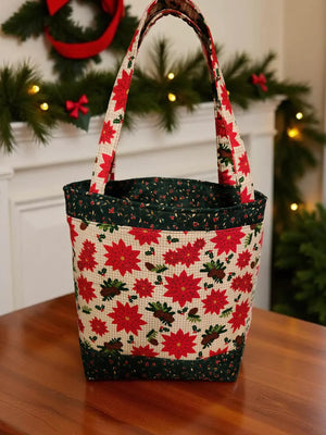 A handbag with a floral pattern of red poinsettias and green leaves sits on a wooden surface, with a wreath and Christmas lights visible in the background.