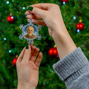A person's hands holding a silver ornament with a picture of a man on it, against a background of a Christmas tree with red and gold ornaments.