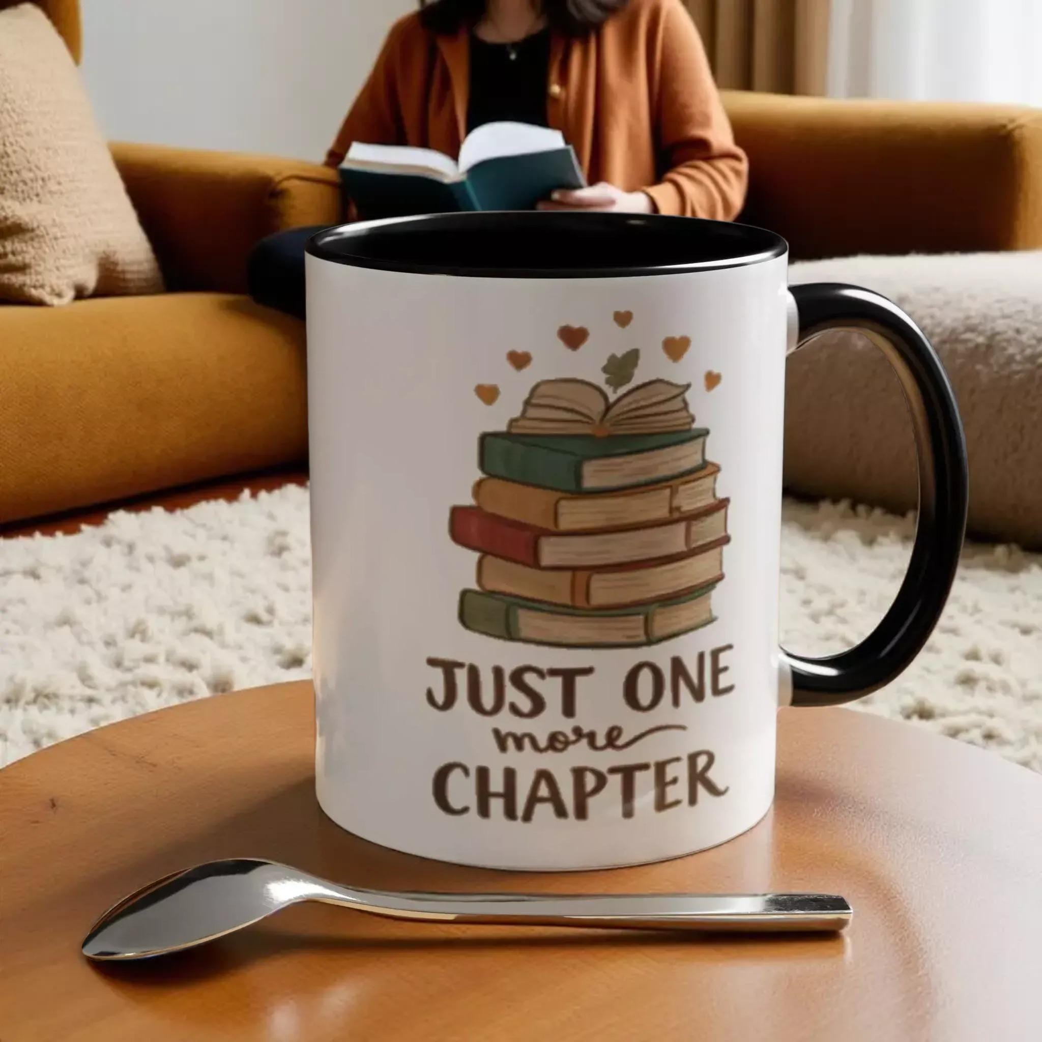 A white mug with a black handle, featuring a stack of books and the text "JUST ONE MORE CHAPTER" printed on it. The mug is placed on a wooden surface, with a spoon nearby.
