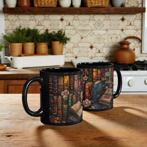 two black mugs with colorful illustrations of books and flowers on them, placed on a desk with a laptop, a corkboard, and some office supplies.