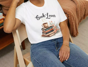A smiling woman wearing a white t-shirt with a graphic of books and a cup of coffee, sitting on a wooden chair.