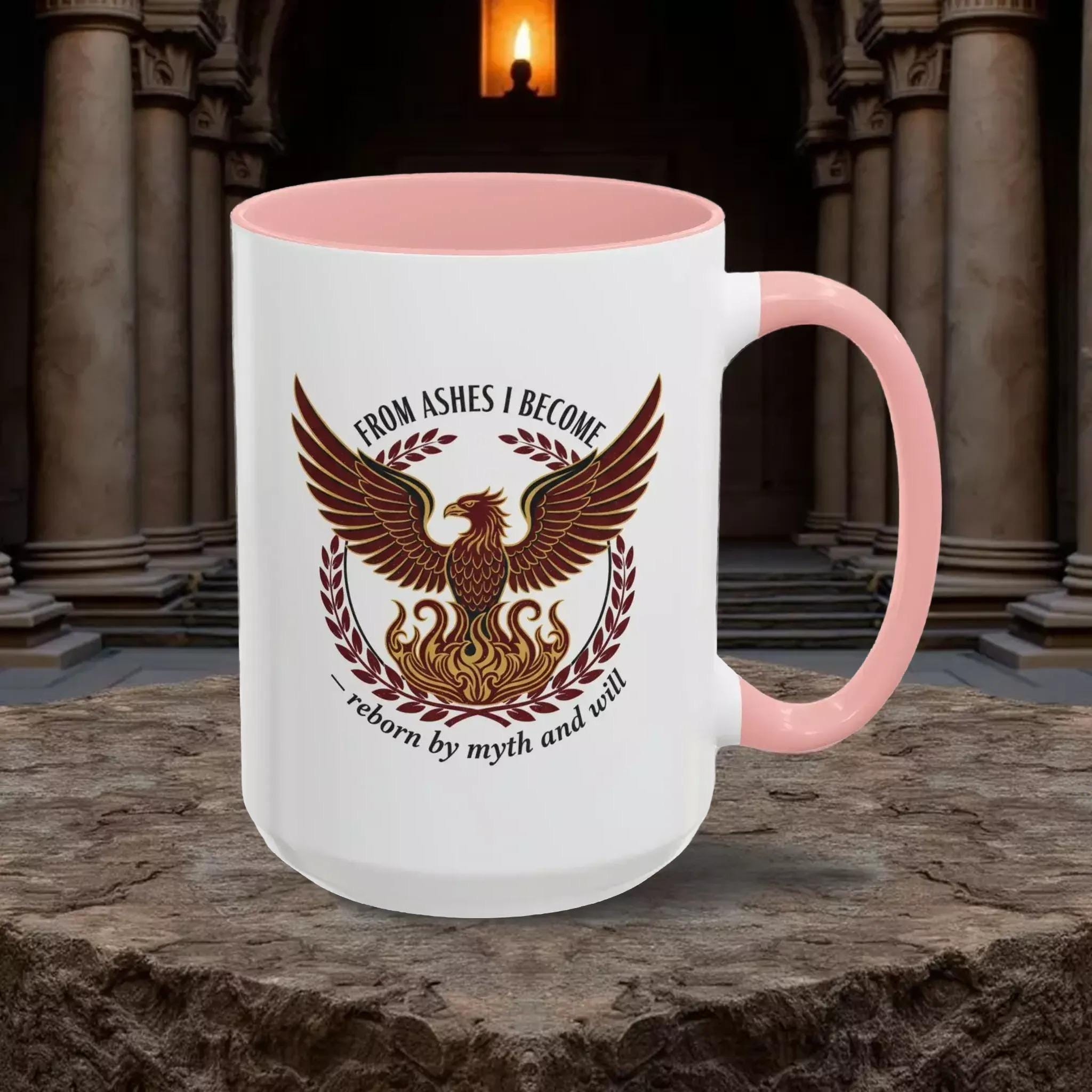 A white mug with a pink interior, featuring a graphic design of a phoenix with wings spread wide, surrounded by laurel branches and flames.