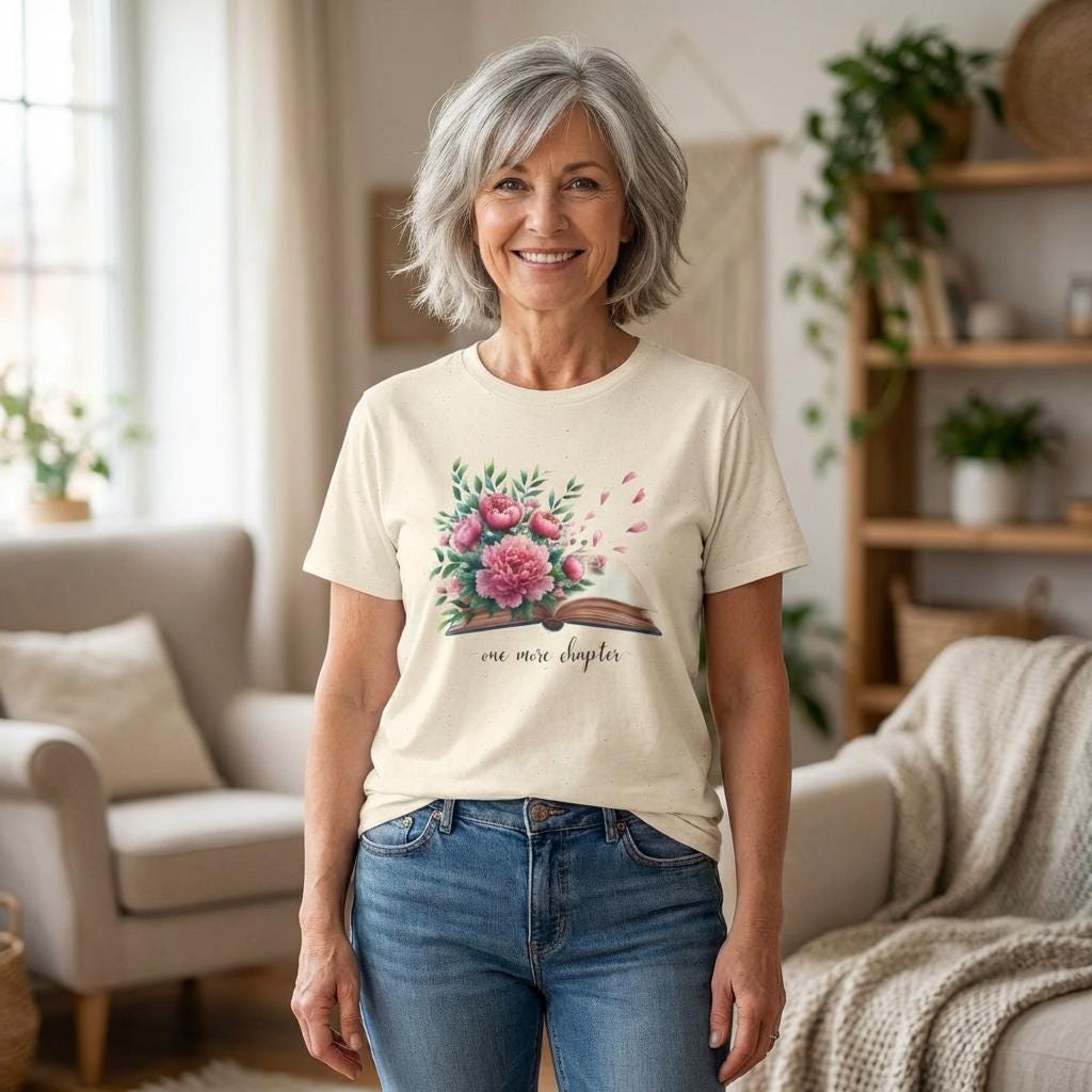 A smiling woman wearing a white t-shirt with a floral design and the text "one more chapter" stands in a living room with a couch and potted plants in the background.