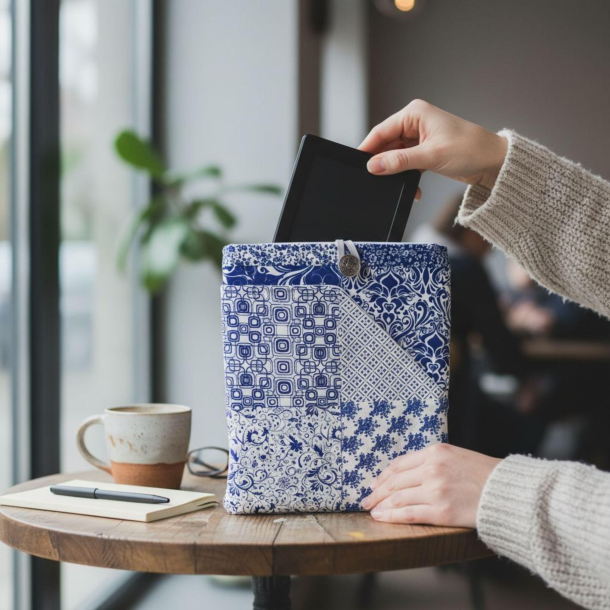 A person's hand is holding a black tablet, which is partially inserted into a blue and white patterned bag, which is placed on a wooden table. There is also a coffee cup and a notebook nearby.