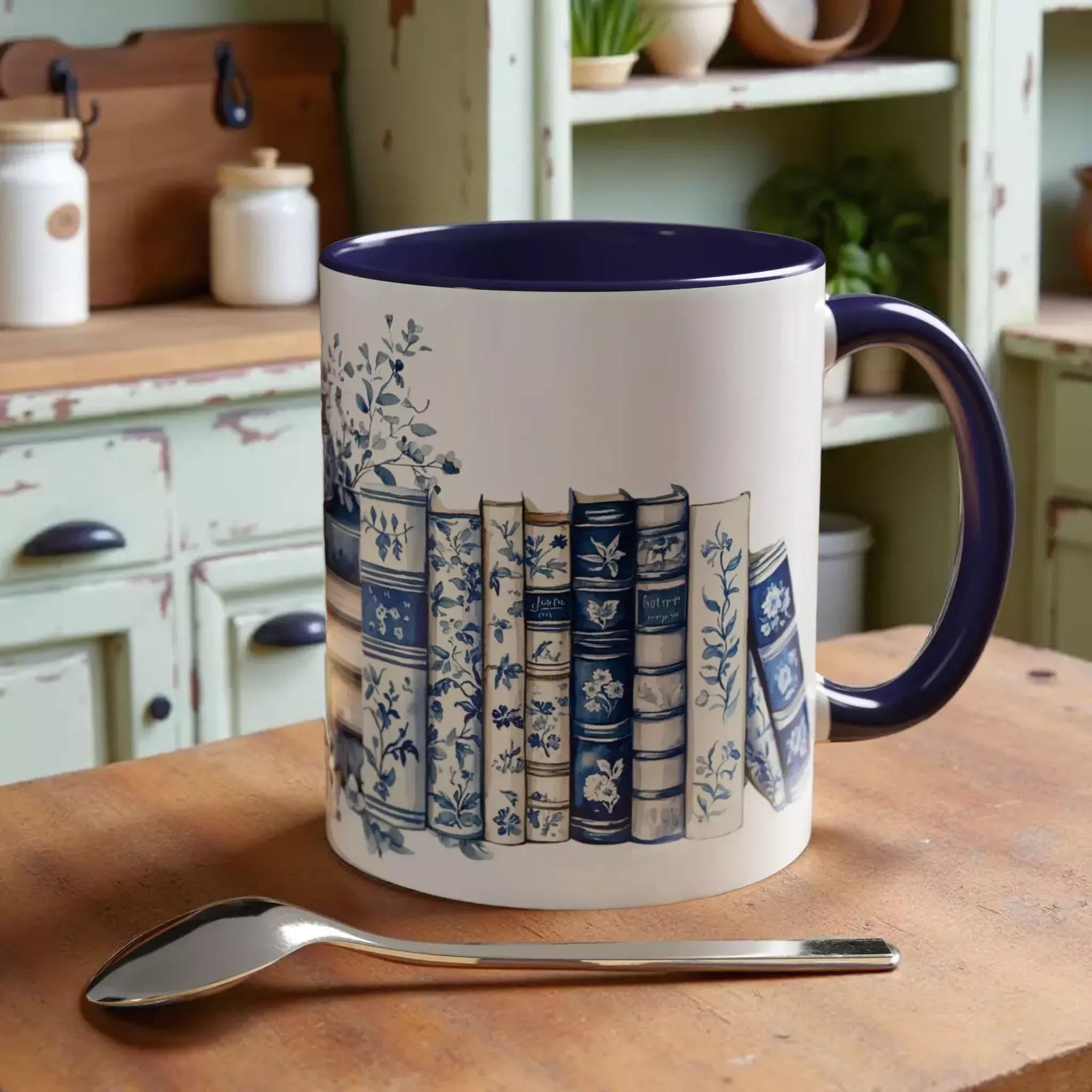 A white mug with a blue rim, featuring a design of blue and white books, is placed on a wooden surface next to a spoon and some coffee beans.