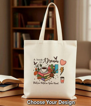 A beige tote bag with a printed design featuring a book, flowers, a cup, and other objects, along with the text "A book dream" and "That you hold in your hand".