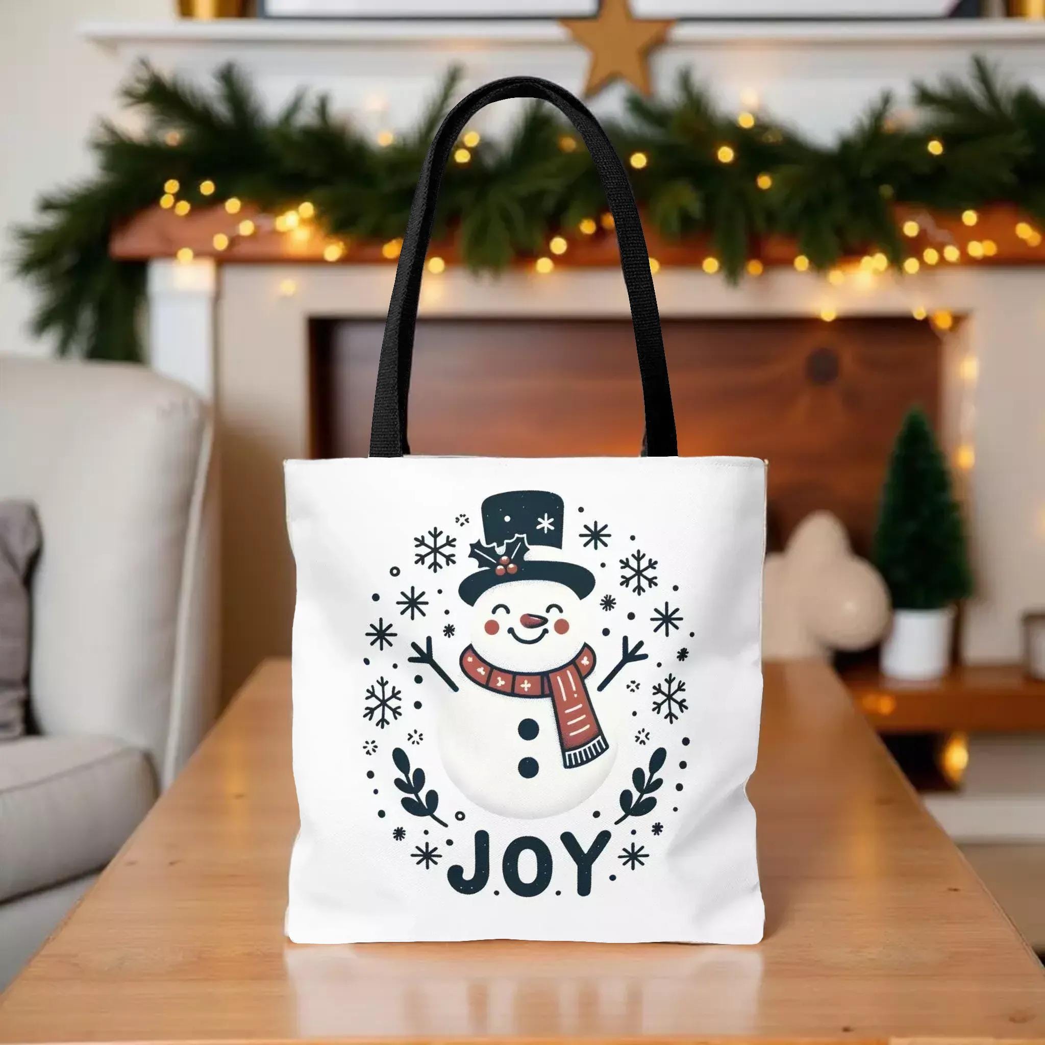 A white tote bag with a snowman design and the word "JOY" printed on it, placed on a wooden surface in front of a fireplace decorated with Christmas lights.