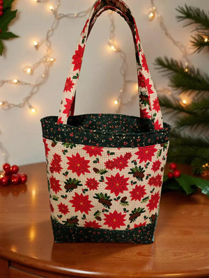 A handbag with a floral pattern and red poinsettia flowers is displayed on a wooden surface, with a Christmas tree and twinkling lights in the background.