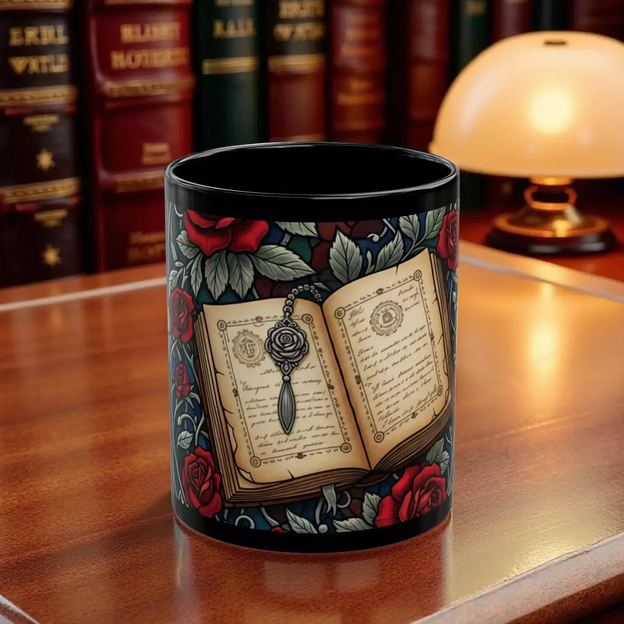 A black mug with a floral design featuring an open book with a rose and a key, surrounded by red roses and leaves.