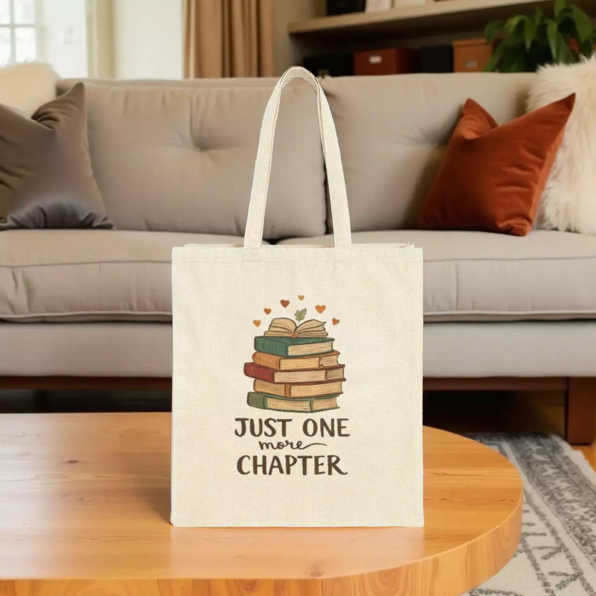 A beige tote bag with a printed design of a stack of books and the text "Just one more chapter" printed on it.
