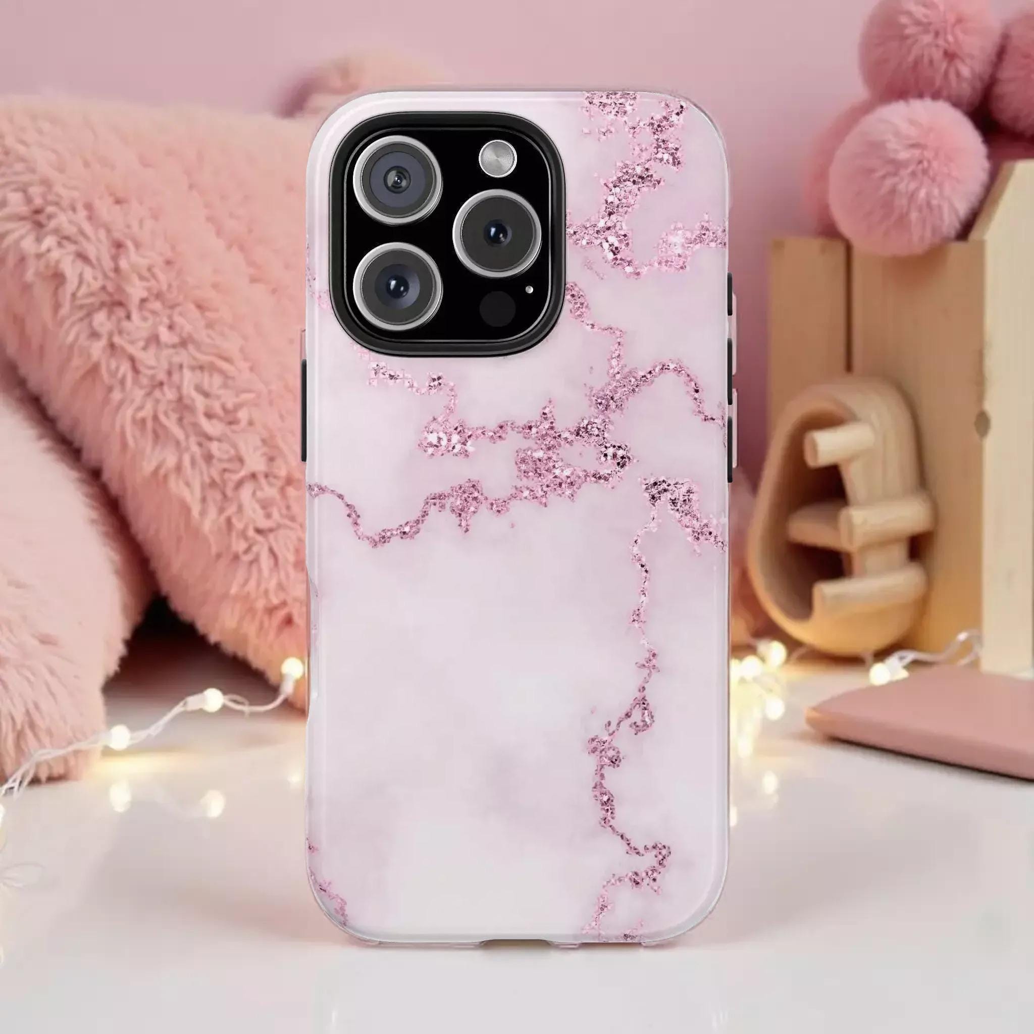 A pink marble-patterned phone case with glittery accents, placed on a white surface with a pink stuffed animal and string lights in the background.