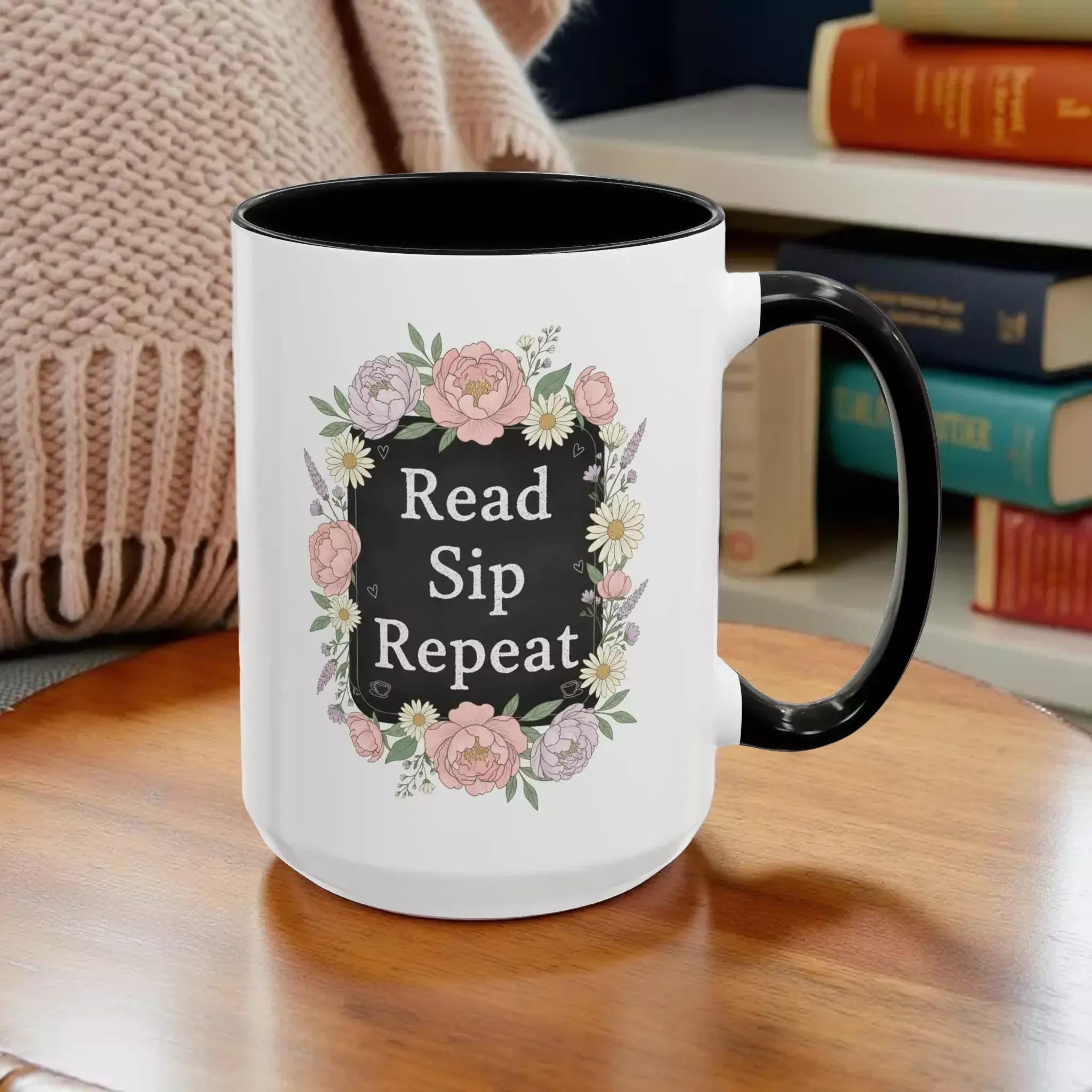 A white mug with a black handle, featuring a floral design and the text "Read, Sip, Repeat" printed on it.