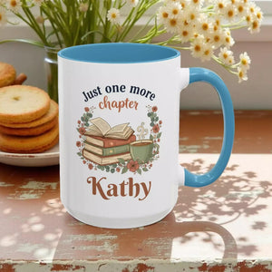 A white mug with a blue handle, featuring a design with a stack of books, a cup of coffee, and the text "Just one more chapter Kathy" printed on it.