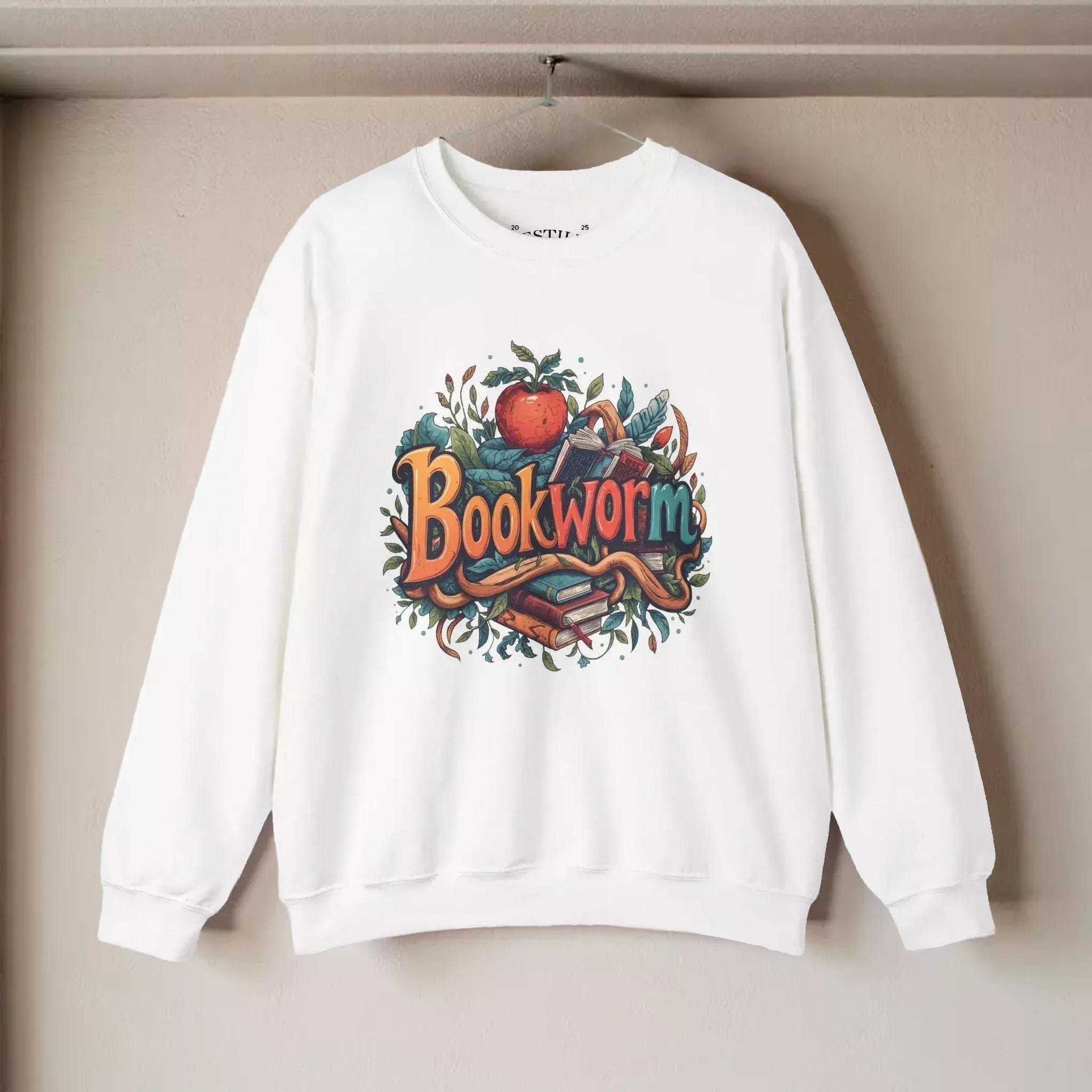 A white sweatshirt with a colorful graphic design featuring the word "Bookworm" surrounded by various objects such as books, leaves, and an apple.