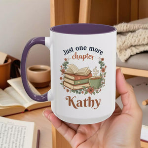 A white mug with a purple handle, featuring an illustration of a stack of books and a cup of tea, along with the text "Just one more chapter Kathy" printed on it.