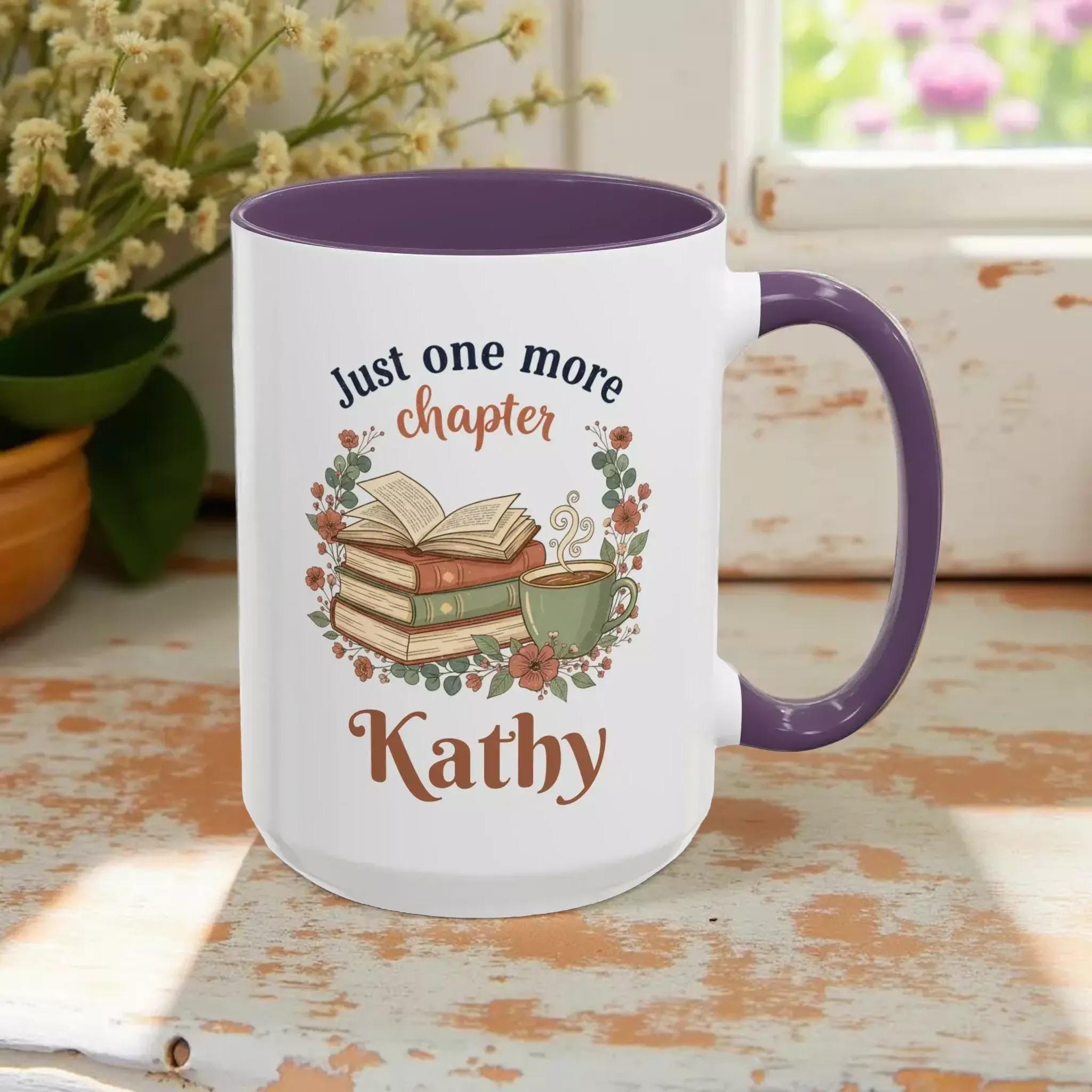 A white mug with a purple handle, featuring a design of a stack of books, a cup of coffee, and the text "Just one more chapter Kathy" printed on it.
