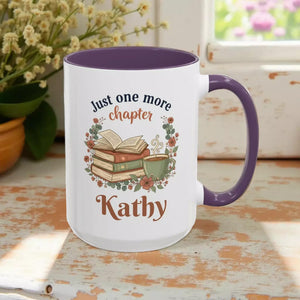 A white mug with a purple handle, featuring a design of a stack of books, a cup of coffee, and the text "Just one more chapter Kathy" printed on it.