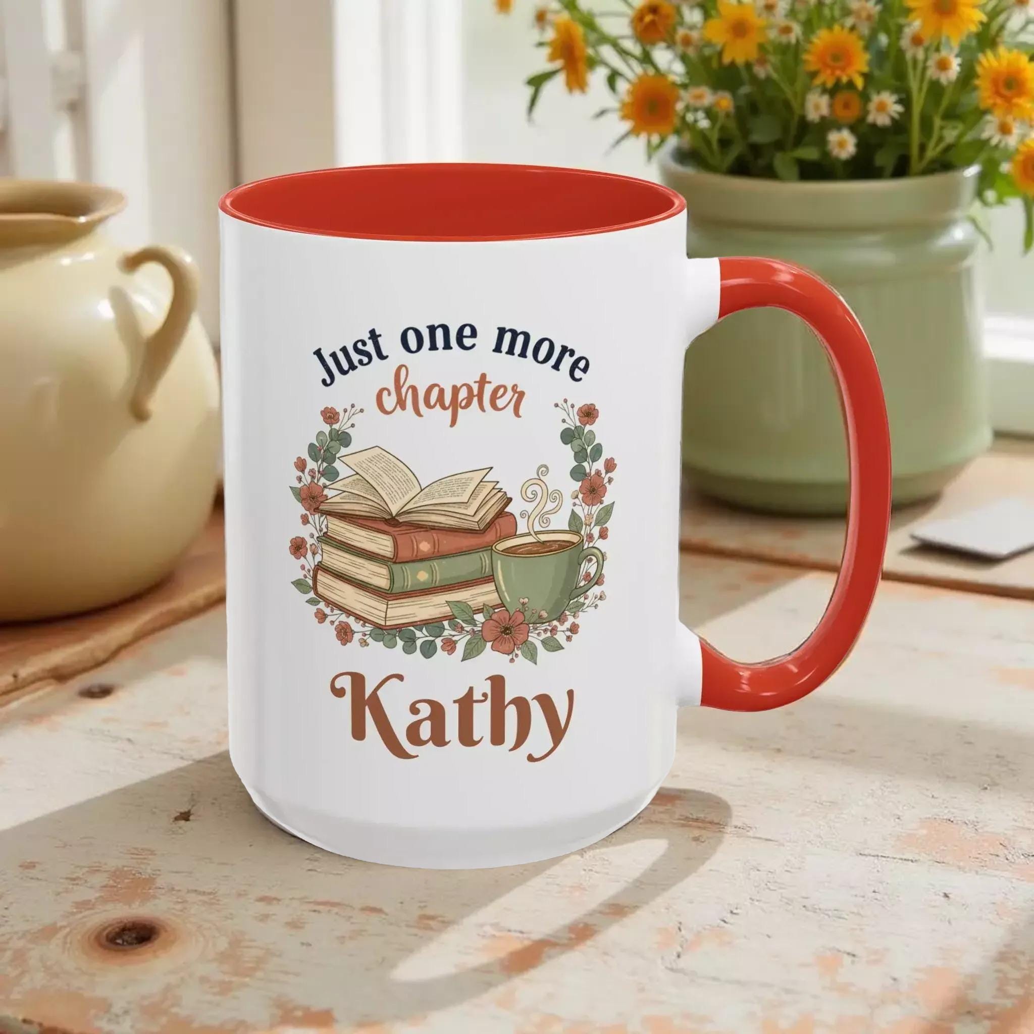 A white mug with a red rim, featuring a design of books, flowers, and a cup of coffee. The text on the mug reads "Just one more chapter Kathy".