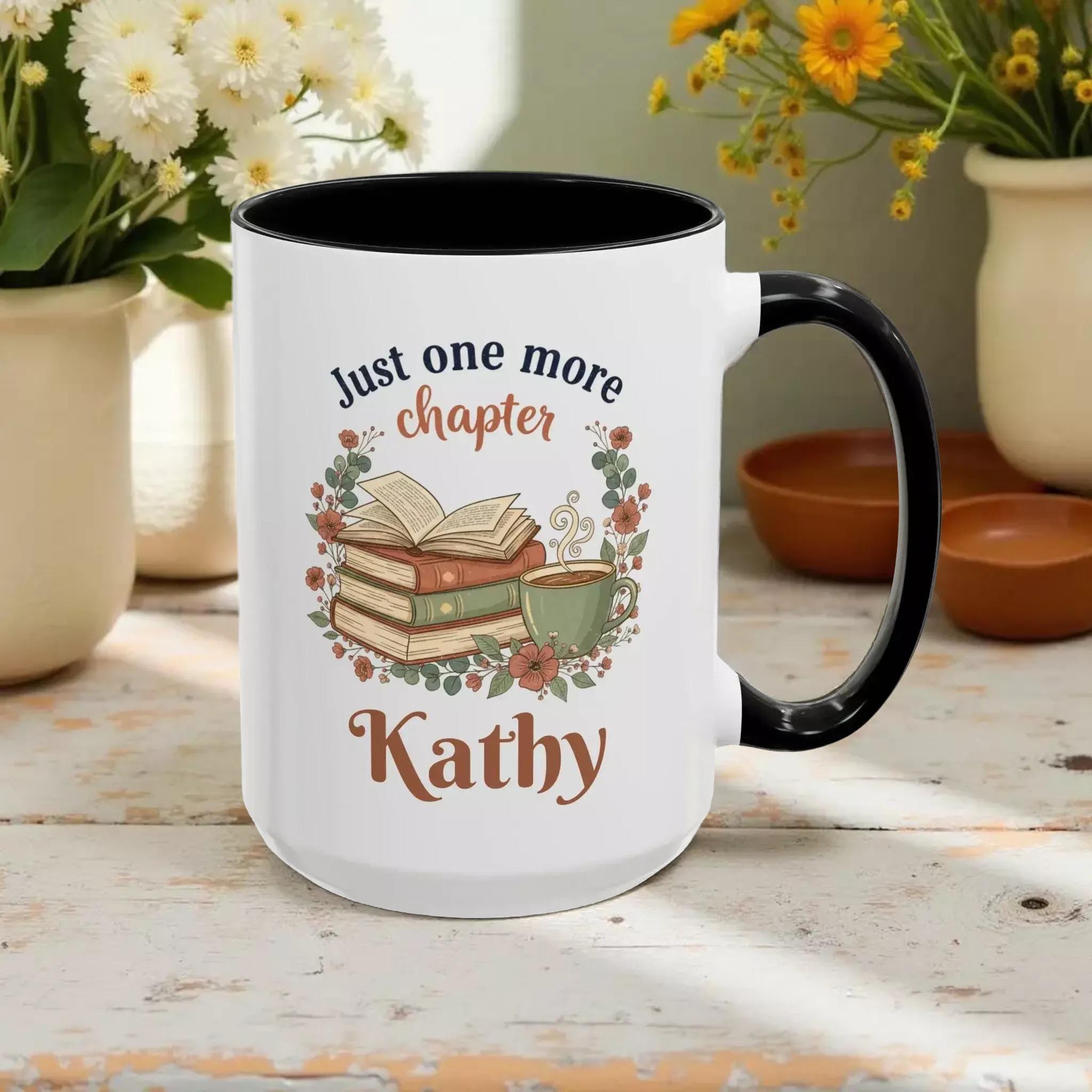 A white mug with a black handle, featuring a design of a stack of books, a cup of coffee, and the text "Just one more chapter Kathy" printed on it.