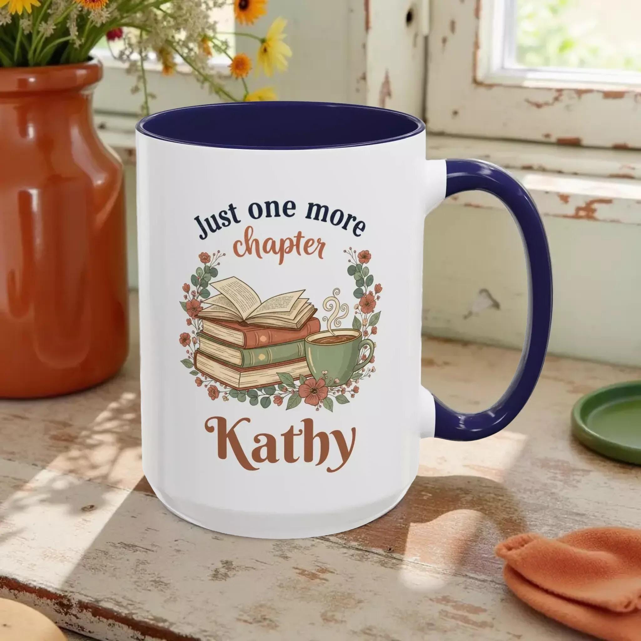 A white mug with a blue rim, featuring a design of a stack of books, a cup of coffee, and the text "Just one more chapter Kathy" printed on it.