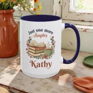 A white mug with a blue rim, featuring a design of a stack of books, a cup of coffee, and the text "Just one more chapter Kathy" printed on it.