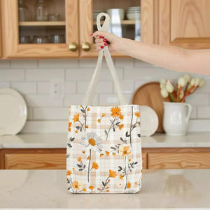 A hand holding a white tote bag with a floral pattern, standing in front of a kitchen counter with various kitchen items visible.