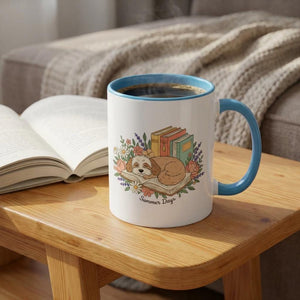 A white mug with a blue handle, featuring a cartoon image of a dog curled up with books, sits on a wooden table next to an open book.