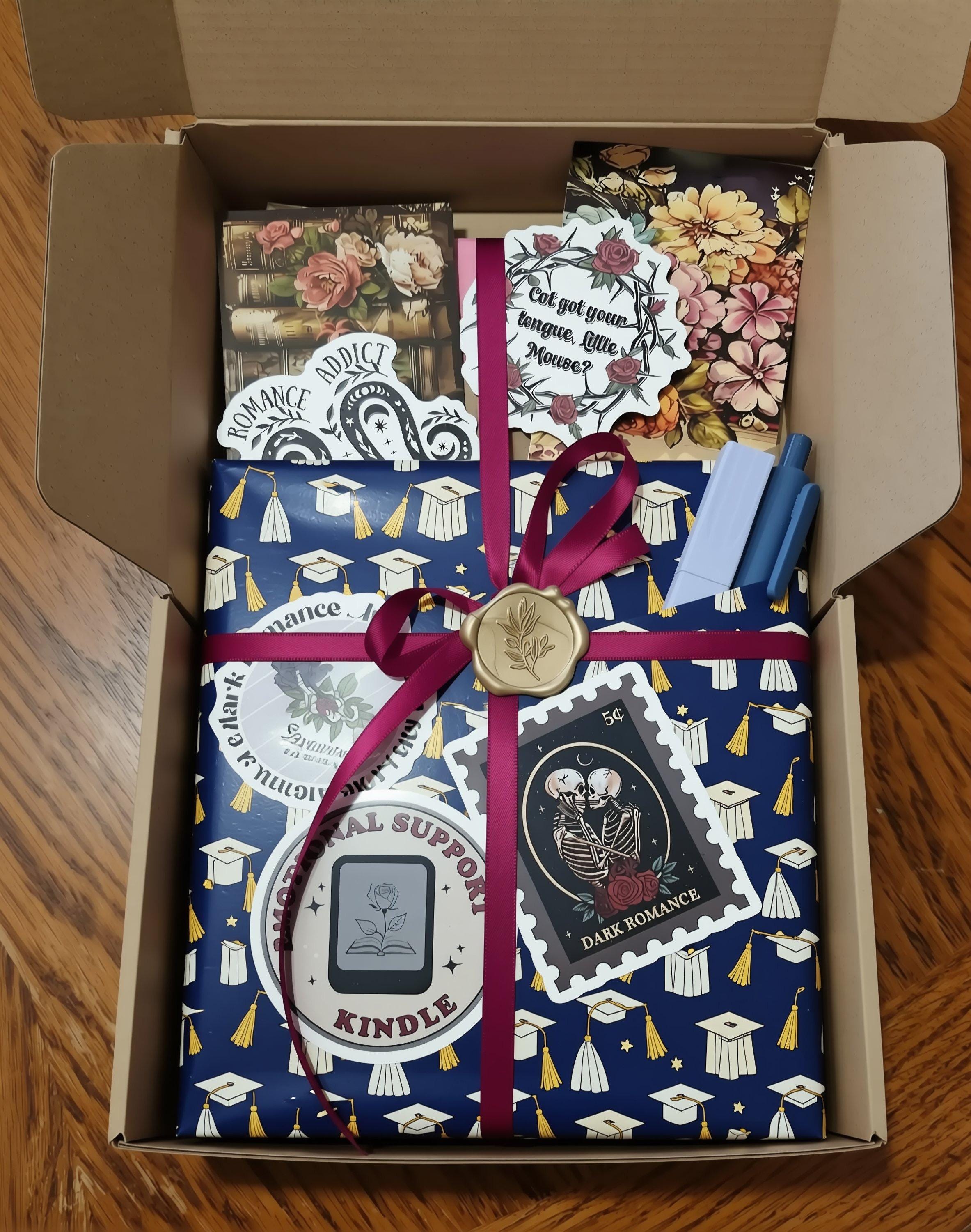 a pink box containing various items, including a book, a card, and some stickers. The box is open, revealing the contents inside.