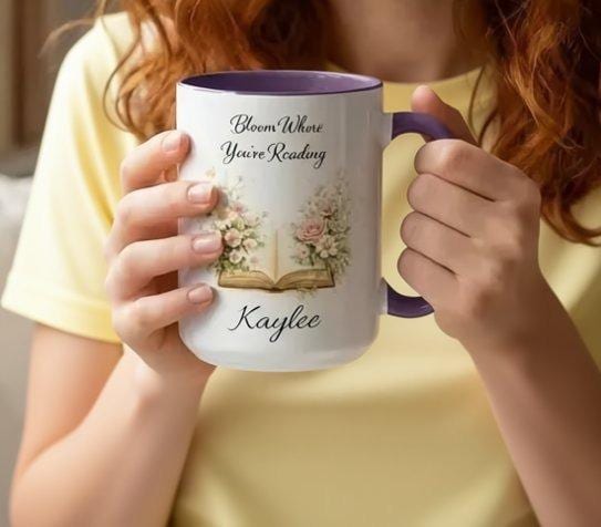 A white mug with a blue handle, featuring an illustration of an open book surrounded by flowers.