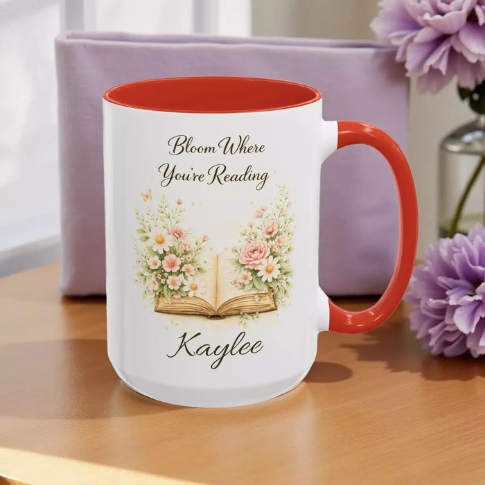 A white mug with a blue handle, featuring an illustration of an open book surrounded by flowers.