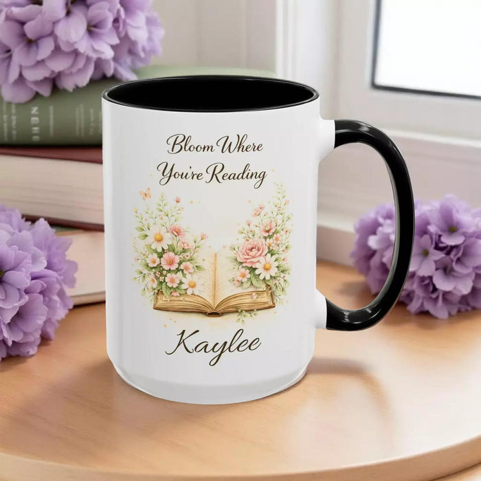 A white mug with a blue handle, featuring an illustration of an open book surrounded by flowers.