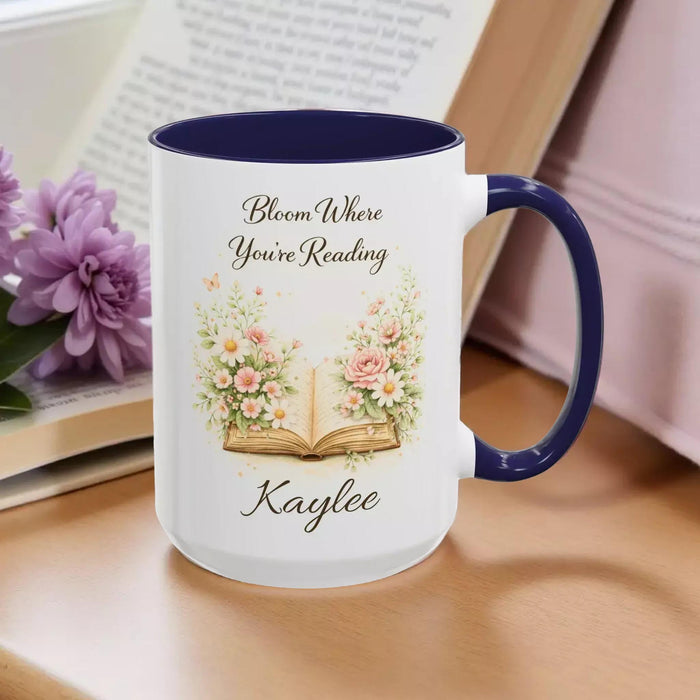 A white mug with a blue handle, featuring an illustration of an open book surrounded by flowers.