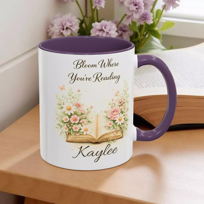 A white mug with a blue handle, featuring an illustration of an open book surrounded by flowers.