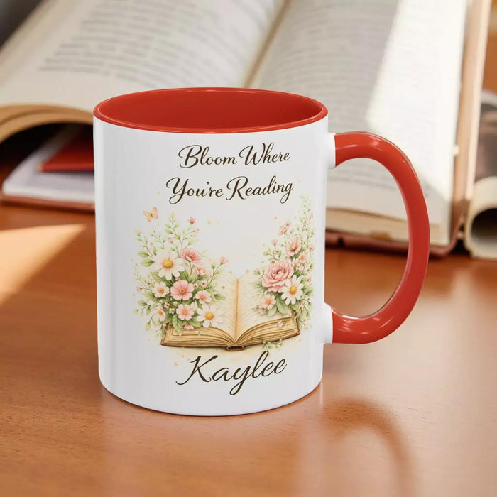 A white mug with a blue handle, featuring an illustration of an open book surrounded by flowers.