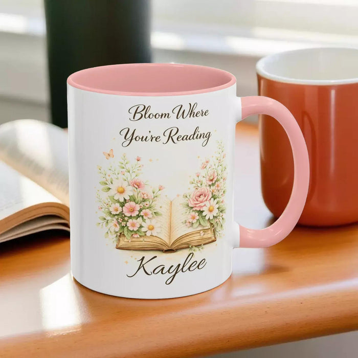 A white mug with a blue handle, featuring an illustration of an open book surrounded by flowers.