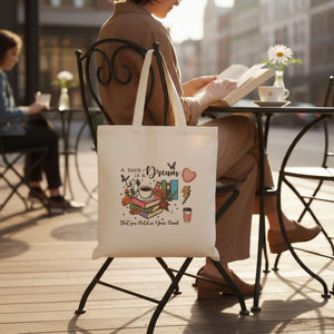 A person is sitting at a table, reading a book, with a white tote bag featuring a book-themed design hanging from the back of a chair.