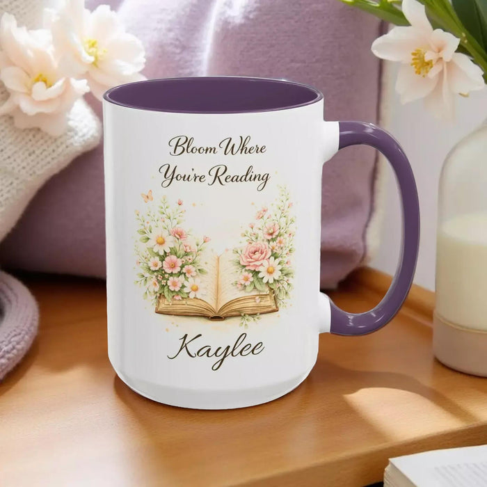 A white mug with a blue handle, featuring an illustration of an open book surrounded by flowers.