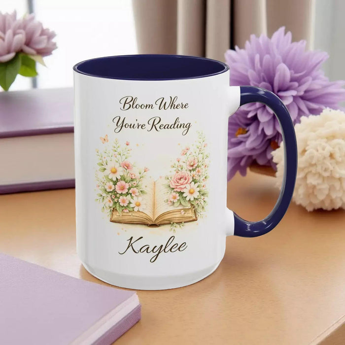 A white mug with a blue handle, featuring an illustration of an open book surrounded by flowers.