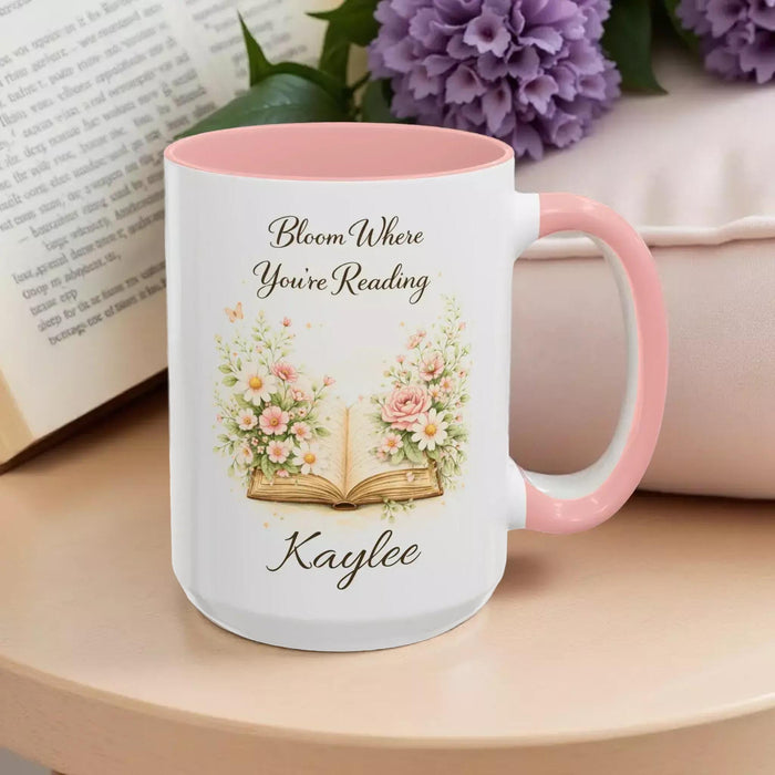 A white mug with a blue handle, featuring an illustration of an open book surrounded by flowers.