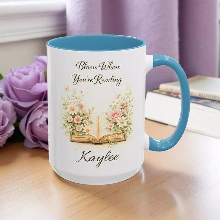 A white mug with a blue handle, featuring an illustration of an open book surrounded by flowers.