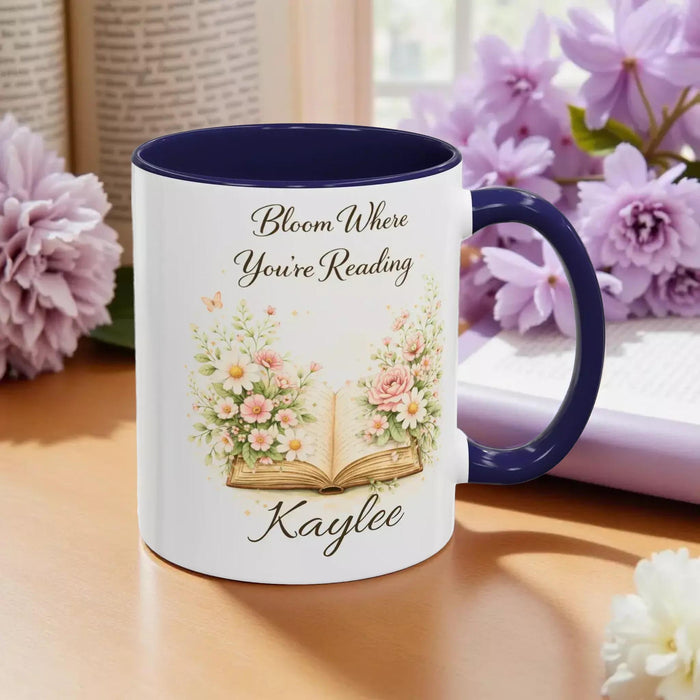 A white mug with a blue handle, featuring an illustration of an open book surrounded by flowers.