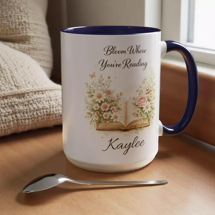 A white mug with a blue handle, featuring an illustration of an open book surrounded by flowers.