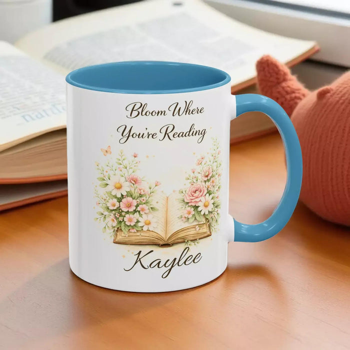 A white mug with a blue handle, featuring an illustration of an open book surrounded by flowers.