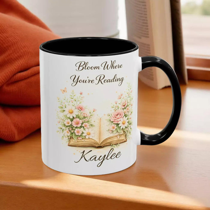 A white mug with a blue handle, featuring an illustration of an open book surrounded by flowers.