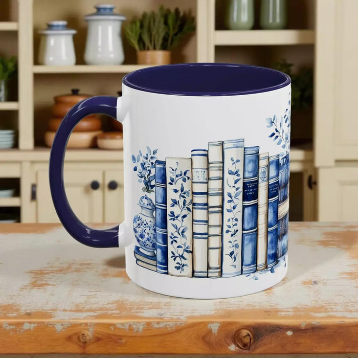 A white tote bag with a blue floral design and a stack of blue and white books, along with a matching blue and white mug, are displayed on a wooden surface.