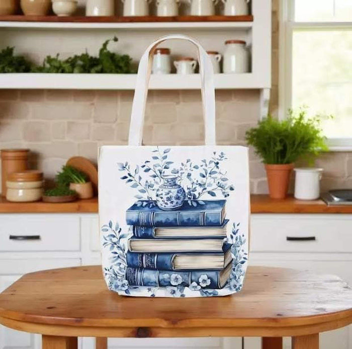 A white tote bag with a blue floral design and a stack of blue and white books, along with a matching blue and white mug, are displayed on a wooden surface.