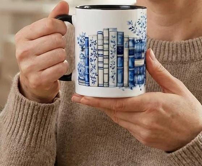 A white tote bag with a blue floral design and a stack of blue and white books, along with a matching blue and white mug, are displayed on a wooden surface.