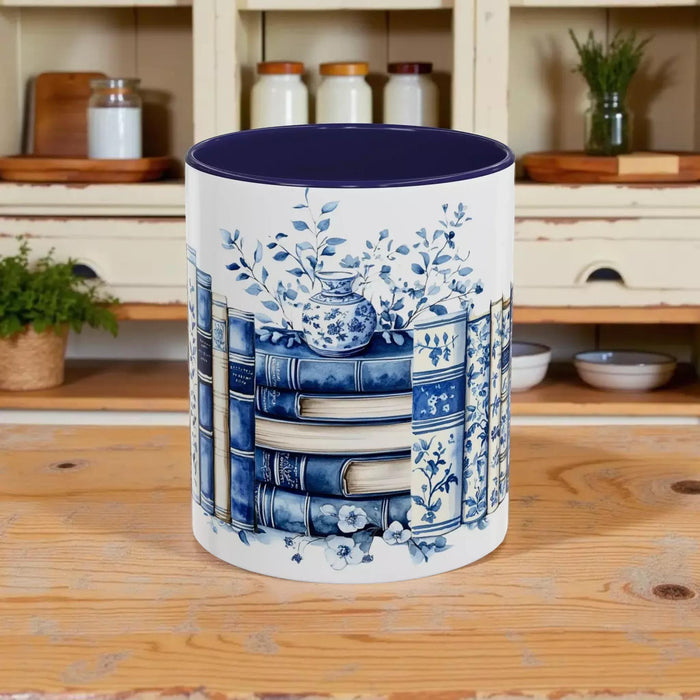 A white tote bag with a blue floral design and a stack of blue and white books, along with a matching blue and white mug, are displayed on a wooden surface.